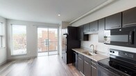 This single-floor unit showcases a thoughtfully designed open-concept layout with contemporary finishes throughout. Wood-style plank flooring flows seamlessly across the living and kitchen areas, contributing to a warm yet modern aesthetic.

The kitchen is a standout feature, equipped with dark espresso cabinetry extending from floor to ceiling, offering generous storage capacity. Light speckled countertops provide a complementary contrast to the dark cabinetry, while black stainless appliances — including a refrigerator, electric range, built-in microwave, and dishwasher — complete the well-appointed space. A single-basin sink with a brushed nickel faucet adds a refined touch.

The open living area is bright and airy, benefiting from multiple windows and a sliding glass door that leads to a private balcony overlooking surrounding rooftops, allowing for natural light and outdoor views.

The bathroom is cleanly finished, featuring dark vanity cabinetry with a speckled countertop, a combination tub and shower with brushed nickel fixtures, and coordinating wood-style flooring. Exposed ductwork visible near the kitchen ceiling adds an industrial design element. Recessed lighting and neutral gray walls maintain a clean, modern atmosphere throughout.