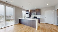 This contemporary single-floor space showcases a thoughtfully designed open-concept layout that seamlessly connects the kitchen and living areas. The kitchen is a standout feature, centered around a large waterfall-edge island with a white quartz countertop and dark wood paneling, complemented by rich, dark-toned cabinetry that extends to the ceiling. Stainless steel appliances — including a refrigerator, electric range, and over-the-range microwave — are paired with a marble-look tile backsplash, adding a refined touch. Pendant lighting above the island and recessed ceiling lights provide ample illumination throughout. Light hardwood flooring flows consistently throughout the space, enhancing its warm, cohesive feel. The living area features a built-in fireplace, serving as a focal point of the room. Large sliding glass doors and windows allow generous natural light to fill the space. The bedrooms are well-proportioned, each featuring the same hardwood flooring, neutral gray walls, and built-in closets with organized shelving. A ceiling fan is present in one of the bedrooms. The bathroom is finished with striking full-height marble-look tile surrounding a walk-in glass shower with a multi-function chrome fixture system, delivering a clean, spa-like aesthetic throughout.