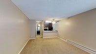 This single-floor studio unit offers an open-concept layout that maximizes the use of its compact footprint. The living area is finished with wall-to-wall carpeting in a neutral beige tone, creating a warm and cohesive atmosphere throughout the space. A ceiling fan is centrally positioned overhead, providing both air circulation and a subtle decorative element. The textured ceiling adds a classic residential touch.

The kitchen is defined by a peninsula-style breakfast bar that separates it from the living area while maintaining an open, connected feel. Dark espresso-toned cabinetry lines the kitchen walls, complemented by black appliances including a refrigerator and range. The contrast between the dark cabinetry and the light-colored walls gives the kitchen a clean, defined look.

A hallway visible from the main living space leads toward the bathroom, which appears to feature a vanity with a mirror and continued dark cabinetry consistent with the kitchen's design. A baseboard heater runs along the exterior wall, providing efficient heating. The neutral wall color throughout ties the space together, offering a versatile backdrop well-suited to a variety of furnishing styles.