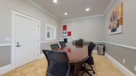 This single-room conference space is thoughtfully designed to support professional meetings in a refined setting. The room centers around a rich, oval-shaped cherry wood conference table surrounded by several ergonomic mesh-back chairs, comfortably accommodating a small group. Travertine tile flooring in warm beige tones runs throughout, lending an upscale, clean aesthetic that complements the neutral gray walls.

Crown molding accents the ceiling perimeter, adding an architectural elegance consistent with the room's polished character. A chair-rail molding runs along the lower portion of the walls, visually dividing the two-toned gray paint scheme and contributing a classic, structured look. Recessed lighting is positioned in the ceiling to provide even, functional illumination.

Natural light enters through multiple windows fitted with white plantation shutters, allowing adjustable light control while offering glimpses of surrounding greenery. A rustic wood console table along one wall provides a surface for decorative items, while building systems—including a fire alarm panel and security controls—are mounted neatly on the adjacent wall. A small utility cart offers additional storage. The overall ambiance is professional yet welcoming, balancing practical functionality with tasteful interior detailing.