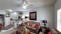 This single-story space showcases an open-concept layout that seamlessly connects the living area and kitchen, creating a cohesive and functional environment. The living area is anchored by a comfortable upholstered sofa accented with decorative pillows in rich red and floral patterns, complemented by a wooden coffee table that adds a warm, traditional touch. A ceiling fan with a built-in light fixture is centrally mounted overhead, providing both airflow and ambient lighting. The textured ceiling adds subtle architectural character to the space. A bold, framed wall art piece featuring a tree motif serves as a prominent focal point on the main wall, enhancing the room's visual appeal. The kitchen, visible from the living area, features light wood cabinetry, a built-in microwave, and a breakfast bar with white bar stools, offering a casual dining option. Under-cabinet and overhead lighting illuminate the kitchen workspace effectively. The neutral wall tones throughout provide a versatile backdrop, while table lamps in both areas contribute to a warm, layered lighting scheme. The overall ambiance is cozy and inviting, blending traditional furnishings with practical, everyday functionality.