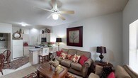 This single-story space showcases an open-concept layout that seamlessly connects the living area and kitchen, creating a cohesive and functional environment. The living area is anchored by a comfortable upholstered sofa accented with decorative pillows in rich red and floral patterns, complemented by a wooden coffee table that adds a warm, traditional touch. A ceiling fan with a built-in light fixture is centrally mounted overhead, providing both airflow and ambient lighting. The textured ceiling adds subtle architectural character to the space. A bold, framed wall art piece featuring a tree motif serves as a prominent focal point on the main wall, enhancing the room's visual appeal. The kitchen, visible from the living area, features light wood cabinetry, a built-in microwave, and a breakfast bar with white bar stools, offering a casual dining option. Under-cabinet and overhead lighting illuminate the kitchen workspace effectively. The neutral wall tones throughout provide a versatile backdrop, while table lamps in both areas contribute to a warm, layered lighting scheme. The overall ambiance is cozy and inviting, blending traditional furnishings with practical, everyday functionality.