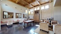 This single-floor clubroom is a thoughtfully designed social space that blends rustic warmth with contemporary comfort. The room immediately impresses with its soaring ceilings adorned with rich, exposed wood beam detailing and polished concrete floors that reflect natural light throughout. Stacked stone accent walls add texture and a sense of architectural character, complementing the warm wood tones found across the space.

At the heart of the room sits a classic billiard table, anchoring the main gathering area and surrounded by ample open floor space. Pendant lighting hangs overhead, providing warm illumination that enhances the inviting atmosphere. A built-in wet bar or beverage station with cabinetry is tucked along one wall, ideal for entertaining.

A built-in banquette seating area lines one wall, featuring neutral upholstery with accent pillows, paired with round café-style tables and chairs — perfect for casual conversation or informal dining. A pair of large decorative mirrors adds depth to this seating nook.

A dedicated media lounge offers a cozy retreat, with a full wall of warm wood paneling framing a built-in entertainment center complete with a flat-screen TV and media storage, flanked by plush upholstered armchairs. Floor-to-ceiling multi-pane glass doors flood the space with natural light and provide views of the outdoors, creating an airy, open ambiance throughout.