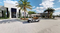 This single-story property presents a polished, resort-inspired exterior characterized by clean, modern architecture with white and neutral-toned facades accented by dark window frames. The entrance area features an expansive paver courtyard, lending a sense of openness and elegance to the approach. Lush tropical landscaping is a defining feature, with tall palm trees, Italian cypress columns, and vibrant flowering ground cover creating a manicured, welcoming atmosphere. A multi-passenger golf cart parked prominently in the courtyard suggests a community-oriented environment, possibly serving as a shuttle or amenity vehicle for residents or guests. In the background, a multi-story residential or mixed-use structure is visible, hinting at a broader development or campus setting. The wide-open paver surface provides ample space for vehicle and pedestrian circulation. Generous natural light and a bright Florida sky enhance the overall ambiance, reinforcing the tropical, upscale character of the property. The well-kept grounds and attention to detail in the landscaping indicate a professionally managed, amenity-rich environment.