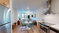 Located on NE Hassalo Street in Portland, Oregon, this one-bedroom, one-bathroom residence offers a thoughtfully designed, single-floor layout with a clean, modern aesthetic throughout. Light hardwood flooring runs continuously across the space, contributing to a warm and cohesive feel.

The open-plan living, dining, and kitchen area forms the heart of the unit. The kitchen is well-appointed with warm wood cabinetry, white quartz countertops, a subway tile backsplash, stainless steel appliances including a range with overhead hood, dishwasher, and full-size refrigerator, along with ample storage in both upper and lower cabinets. A dedicated dining area sits adjacent, offering comfortable space for a table and chairs. The living room features large windows with wood-framed surrounds that bring in generous natural light and frame views of the surrounding Portland cityscape.

The bedroom is a quiet, well-proportioned retreat with its own large window overlooking greenery and neighboring buildings, plus a closet for storage. The bathroom is clean and functional, featuring a wood-toned vanity with white countertop, a full tub-shower combination with tiled surround, and wood-look flooring. A welcoming entrance hall and additional closet storage round out this efficiently organized and stylish urban residence.