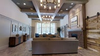 This single-floor space makes an immediate impression with its dramatically high coffered ceilings, which are accented by recessed lighting and two substantial industrial-style chandeliers that serve as the room's focal points. The ceiling's architectural detailing adds depth and visual interest, framing the open layout with a refined, structured elegance.

The main living area is generously proportioned, accommodating a large sectional sofa and coordinating blue accent chairs arranged to encourage both conversation and relaxation. A stone fireplace anchors one wall, providing a warm, traditional counterpoint to the otherwise contemporary design. Adjacent to the fireplace, built-in cabinetry and panel molding add architectural character.

Large windows along the rear wall flood the space with natural light, offering views of lush greenery beyond. Sheer white curtains frame the windows softly, balancing privacy with brightness. The neutral color palette of warm grays and whites is carried throughout, with wainscoting along the lower walls contributing a classic, polished finish. The overall ambiance is welcoming and upscale, blending traditional and transitional design elements seamlessly.