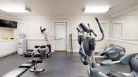 Located at 1250 5th St NE in Hickory, NC, this single-floor fitness center offers a well-equipped and neatly organized workout space. The main exercise room features warm beige walls accented by crisp white crown molding and chair rail trim, lending a polished, finished look to the facility. Durable rubber flooring covers the workout area, providing both safety and practicality for high-traffic use. A large wall mirror spans a significant portion of one wall, visually expanding the space and allowing users to monitor their form during exercise. The room is furnished with multiple cardio machines, including treadmills, elliptical trainers, a stationary bike, and a multi-use strength training station, making it a versatile option for a range of fitness routines. Overhead flush-mount lighting ensures the space is well-lit throughout. Natural light filters in through windows fitted with blinds, adding brightness without sacrificing privacy. An adjoining restroom provides added convenience, featuring a toilet, sink with vanity, window with blinds, and speckled tile-style flooring. A fire extinguisher is mounted near the restroom entrance, reflecting attention to safety. This functional fitness space is well-suited for a community facility, residential complex, or small commercial gym setting.