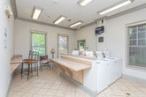 This shared laundry facility is designed with both functionality and comfort in mind. The room features a row of modern front-loading washers and dryers neatly arranged along one wall, providing ample capacity for residents. A generous laminate folding counter wraps along the center of the space, offering plenty of room for sorting and folding laundry. The neutral-toned walls are complemented by crown molding details near the ceiling, lending the room a polished, well-maintained appearance. Fluorescent ceiling fixtures provide consistent, bright overhead lighting throughout. Multiple windows fitted with horizontal blinds allow natural light to filter in, creating an inviting atmosphere with views of the surrounding greenery outside. A wall-mounted flat-screen television is positioned for easy viewing, adding a leisure element while residents wait. Seating is available near the windows, making the wait more comfortable. The space is clean, organized, and thoughtfully laid out to accommodate multiple users simultaneously, reflecting a practical and user-friendly design approach.
