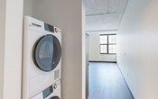 Located at 401 E State St in downtown Ithaca, NY, this 1-bedroom, 1-bathroom unit showcases a modern loft-inspired aesthetic through its open layout and industrial design elements. The living and kitchen area serves as the heart of the unit, featuring light wood-tone flooring, exposed ceiling beams, and visible piping that contribute to its industrial character. Large multi-pane windows flood the space with natural light, enhancing the airy feel of the high ceilings. A wall-mounted mini-split system provides climate control throughout.

The kitchen is outfitted with slate-blue cabinetry paired with white countertops, a subway tile backsplash, and a curved kitchen island with a ribbed pedestal base. A full-size refrigerator and overhead pendant lighting complete the well-appointed cooking space.

A dedicated closet houses a stacked washer and dryer unit, offering convenient in-unit laundry. Two closets provide ample storage throughout the unit. The bathroom features a gray vanity with black fixtures, a framed mirror, and a walk-in glass-enclosed shower with large-format gray tile detailing. The overall design balances contemporary finishes with industrial charm, creating a cohesive and functional living environment.
