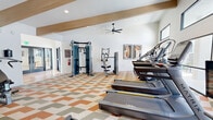 This 770 square foot fitness center, located at 699 W Magee Rd in Tucson, AZ, is a state-of-the-art facility designed to cater to various exercise needs. The space features high ceilings with exposed wooden beams and recessed lighting, creating an open and airy atmosphere. Large windows allow natural light to flood the room, enhancing the bright and energizing environment.

The facility is equipped with a wide range of modern exercise machines, including treadmills, ellipticals, stationary bikes, and weight training equipment. A dedicated area for free weights and resistance training is visible, complete with weight racks and benches. The layout is thoughtfully designed to optimize space usage while ensuring ample room for movement between equipment.

One of the most striking features is the vibrant geometric patterned flooring in shades of orange, beige, and olive green, which adds a dynamic visual element to the space. This flooring not only enhances the aesthetic appeal but also provides a durable surface suitable for heavy gym equipment.

The center is outfitted with amenities such as ceiling fans for air circulation, strategically placed mirrors to assist with form during workouts, and what appears to be a water station for hydration. Safety features like a fire extinguisher are also visible, ensuring a secure exercise environment.