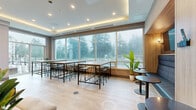 Located at 1915 Sooke Rd in Colwood, BC, this well-appointed amenity space offers a thoughtfully designed single-floor layout that balances collaborative and private functions. The open lounge and dining area is the heart of the space, featuring light hardwood flooring, tray ceilings with warm indirect LED lighting, and floor-to-ceiling windows that flood the interior with natural light and frame views of the surrounding evergreen treeline. Comfortable upholstered seating is arranged alongside bar-height communal tables and bistro-style seating, creating a versatile environment for both relaxation and casual meetings. A wall-mounted television enhances the lounge area, while a compact kitchenette with wood cabinetry and a sink provides practical convenience. Four sleek, glass-enclosed private office pods lined with black metal framing offer sound-separated spaces ideal for focused work or private calls, each furnished with a desk and seating. The neutral warm-toned palette, clean architectural lines, and mix of natural wood accents with modern materials give the space a polished yet welcoming atmosphere, making it a functional and refined shared amenity.