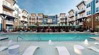 Located at 3908 Independence Blvd in Wilmington, North Carolina, this expansive outdoor amenity space offers a resort-inspired atmosphere designed for relaxation and socializing. The centerpiece is a large, shimmering swimming pool surrounded by rows of chaise lounges and teal-accented umbrellas, creating a vibrant and inviting atmosphere under the open sky. Adjacent to the pool, a bocce ball court adds a recreational element, thoughtfully integrated into the broader courtyard layout.

The hardscaped courtyard features elegant herringbone-patterned brick pavers and large-format stone tiles, providing a polished yet approachable outdoor environment. Tall, mature palm trees are strategically planted throughout the space, lending a coastal, tropical feel that complements Wilmington's warm climate. String lights are strung between poles, setting the stage for evening gatherings and events.

Multiple seating zones are distributed throughout the space, including a covered lounge area furnished with plush sectional sofas accented with teal and blue throw pillows, ideal for casual conversation. A dedicated fire pit area with Adirondack chairs provides a cozy gathering spot. The striking multi-story building facade, clad in bold navy blue and white with brick accents and private balconies, serves as a dramatic backdrop to the entire outdoor living environment.