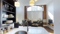 This single-floor space artfully combines historic industrial character with refined modern design. The open-concept layout seamlessly connects the kitchen and living areas, creating a cohesive and inviting environment throughout.

The kitchen is anchored by a generous white quartz countertop that provides ample workspace, complemented by a sleek gooseneck faucet and an undermount sink. Pendant drum lighting overhead adds a warm, contemporary touch.

The living area is the architectural centerpiece of the space, showcasing dramatic exposed brick columns that frame a striking arched window. The window soars toward the high ceiling, flooding the room with natural light and offering views of the surrounding tree canopy. Vertical blinds allow for adjustable privacy and light control.

The living space is thoughtfully arranged with a plush gray sofa, coordinating armchairs, a round wooden coffee table, and layered area rugs that define the seating zone. Flanking end tables with table lamps contribute to the warm, layered ambiance.

The combination of soaring ceilings, raw brick textures, arched architectural detailing, and polished modern furnishings gives the space a distinctive character that balances heritage craftsmanship with contemporary comfort.