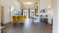 Located at 1901–1931 Hanahan Rd in North Charleston, SC, this single-floor community space is thoughtfully designed to combine function with bold, eclectic style. The main lobby area immediately impresses with its soaring vaulted ceilings clad in white shiplap paneling, large steel-framed glass entry doors, and an abundance of natural light streaming through oversized windows. Dark slate-tiled flooring grounds the space while pendant lighting—including geometric and dome-style fixtures—adds warmth and visual interest overhead.

A central reception counter with a sleek white finish serves as the focal point of the lobby, flanked by a beverage and service bar with gray cabinetry and under-counter refrigeration. Multiple flat-screen monitors are mounted throughout for informational display. The lounge areas feature a curated mix of furnishings, including plush mustard-yellow wingback chairs, brown leather accent chairs with bold yellow throw pillows, and a vintage-style area rug in warm tones. Woven rattan seating and marble-top tables add texture and sophistication.

A distinct teal-walled nook provides an intimate seating area with rattan bistro chairs, a marble pedestal table, and framed botanical artwork, offering a charming contrast to the open main hall. The overall ambiance is vibrant, welcoming, and design-forward, reflecting a carefully curated aesthetic suited to a modern residential community.