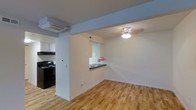 This single-floor space offers a clean and functional layout designed for comfortable everyday living. The main living area features warm hardwood flooring that extends throughout, contributing to a bright and welcoming ambiance. A ceiling fan with an integrated light fixture provides both ventilation and illumination, enhancing the practicality of the space.

A defining architectural detail is the pass-through window connecting the kitchen to the living area, topped with a dark countertop ledge that serves as a convenient bar or serving surface. This design element maintains an open feel while subtly delineating the two spaces.

The kitchen is equipped with black appliances, including a freestanding range, and features white cabinetry overhead, creating a classic contrast. A wall-mounted mini-split air conditioning unit is positioned near the kitchen, ensuring efficient climate control throughout the space.

The walls are finished in crisp white, maximizing the sense of light and openness. Recessed ventilation and clean baseboard trim throughout reflect a well-maintained interior. The overall design balances simplicity and practicality, making the space versatile and move-in ready.