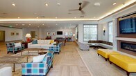 This expansive single-floor amenity space is thoughtfully designed to accommodate a variety of social and recreational activities within one open, flowing layout. Light wood-look tile flooring laid in a herringbone pattern runs throughout, lending warmth and visual continuity across the entire space.

The lounge area is divided into several distinct yet connected zones. A media and fireplace nook anchors one end of the room, featuring a large wall-mounted television framed by a textured tile surround and a sleek linear fireplace set beneath a marble-accented facade. Bold yellow tufted ottomans and a built-in banquette with striped upholstery provide comfortable seating in this area. Adjacent to this, a generous open lounge offers multiple seating arrangements with sofas, plaid accent chairs, and side tables styled in a contemporary palette of teal, yellow, and neutral tones.

A billiards table and a dining or game table with red chairs occupy a central recreational zone, accompanied by a wall-mounted television nearby. The kitchenette area is well-appointed with warm wood cabinetry, dark granite countertops, a sink, and a striking blue mosaic tile backsplash. Pendant lighting with teal accents and recessed cove lighting in the tray ceiling add a polished finishing touch. Large windows and glass doors allow natural light to fill the space.