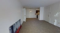 This single-floor space features an open-concept layout that connects the main living area to the kitchen, creating a sense of openness and flow. The living area is finished with wall-to-wall carpet in a neutral tone, providing a comfortable underfoot feel throughout the space. White walls and a smooth white ceiling keep the interior bright and clean, contributing to an airy atmosphere.

A partial wall divides the living area from the kitchen zone, which is visible in the background. The kitchen is equipped with dark wood cabinetry, an overhead range hood, and standard appliances including what appears to be a dishwasher and range. A pendant light hangs above, providing focused task lighting over the kitchen area.

Two doorways are visible — one leading to an adjacent room or hallway on the left side, and another on the right, suggesting additional rooms or utility spaces beyond the main living area. The overall layout is practical and efficient, with a straightforward design that emphasizes functionality. The neutral color palette and open arrangement make the space adaptable to a variety of furniture configurations and uses.