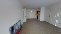 This single-floor space features an open-concept layout that connects the main living area to the kitchen, creating a sense of openness and flow. The living area is finished with wall-to-wall carpet in a neutral tone, providing a comfortable underfoot feel throughout the space. White walls and a smooth white ceiling keep the interior bright and clean, contributing to an airy atmosphere.

A partial wall divides the living area from the kitchen zone, which is visible in the background. The kitchen is equipped with dark wood cabinetry, an overhead range hood, and standard appliances including what appears to be a dishwasher and range. A pendant light hangs above, providing focused task lighting over the kitchen area.

Two doorways are visible — one leading to an adjacent room or hallway on the left side, and another on the right, suggesting additional rooms or utility spaces beyond the main living area. The overall layout is practical and efficient, with a straightforward design that emphasizes functionality. The neutral color palette and open arrangement make the space adaptable to a variety of furniture configurations and uses.
