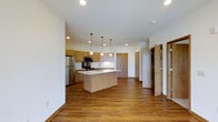 This single-floor space showcases a thoughtfully designed open-concept layout that seamlessly blends the kitchen and living areas into one cohesive, welcoming environment. The kitchen is a standout feature, outfitted with warm-toned wood cabinetry that extends to the ceiling, stainless steel appliances including a refrigerator, microwave, and range, and a sleek central island complete with a sink and pendant lighting above. The pendant lights add a refined, modern touch that complements the recessed lighting throughout the space. Light-toned countertops provide a clean contrast to the cabinetry, while durable hardwood-style flooring in a rich warm hue runs throughout, adding continuity and warmth. The open floor plan allows for generous sightlines across the living area, creating a sense of spaciousness and flow. Crisp white walls and a smooth ceiling contribute to a bright, airy ambiance. Additional doors visible in the background hint at private rooms beyond, suggesting a well-organized and functional floor plan. Overall, the space strikes a balance between contemporary design and everyday practicality, making it an inviting and versatile environment.