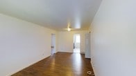 This single-floor space offers a straightforward and functional layout designed for comfortable everyday living. The main living area immediately draws attention with its rich, dark hardwood floors that extend throughout, providing warmth and a classic aesthetic against the crisp white walls and ceiling. The open feel of the space is enhanced by a simple flush ceiling light fixture, keeping the area bright and uncluttered. White baseboards provide a clean finish along the perimeter of the room. Multiple doorways connect the main living area to adjoining rooms, allowing for easy flow throughout the space. One passageway leads to an additional room with matching hardwood flooring, while another opens to a brighter area with a window that brings in natural light — this secondary space appears to include utility features such as a washer or dryer. A classic paneled door on one side of the main room adds a traditional architectural touch. A floor vent is also visible, indicating a built-in heating and cooling system. Overall, the space presents a clean, neutral palette and practical layout that serves as a versatile foundation for a variety of uses.