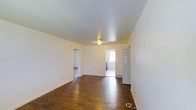 This single-floor space offers a straightforward and functional layout designed for comfortable everyday living. The main living area immediately draws attention with its rich, dark hardwood floors that extend throughout, providing warmth and a classic aesthetic against the crisp white walls and ceiling. The open feel of the space is enhanced by a simple flush ceiling light fixture, keeping the area bright and uncluttered. White baseboards provide a clean finish along the perimeter of the room. Multiple doorways connect the main living area to adjoining rooms, allowing for easy flow throughout the space. One passageway leads to an additional room with matching hardwood flooring, while another opens to a brighter area with a window that brings in natural light — this secondary space appears to include utility features such as a washer or dryer. A classic paneled door on one side of the main room adds a traditional architectural touch. A floor vent is also visible, indicating a built-in heating and cooling system. Overall, the space presents a clean, neutral palette and practical layout that serves as a versatile foundation for a variety of uses.