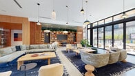 This thoughtfully designed communal space combines a grand lobby with a series of interconnected amenity areas, all unified by a cohesive, contemporary aesthetic. Polished terrazzo flooring extends throughout, reflecting natural light and lending an open, airy quality to every zone.

The entry lobby commands immediate attention with its soaring double-height ceiling, floor-to-ceiling glazing, and a monumental staircase framed by warm wood paneling. Curated artwork and lush potted plants punctuate the space, adding warmth and visual interest alongside a reception desk finished in natural wood tones.

Moving deeper into the space, a generously scaled lounge area features an expansive built-in shelving wall lined with books, décor, and trailing greenery set against a deep teal-painted ceiling. Varied seating arrangements—including plush sofas, sculptural lounge chairs, and a tufted daybed—invite both social gatherings and quiet relaxation. Pendant lighting in mixed styles adds character overhead, while large glazed doors offer views of an outdoor pool area.

A dedicated kitchen and gathering zone includes warm wood cabinetry, black countertops, a decorative patterned tile backsplash, open shelving adorned with plants, and a central bar-height communal table—ideal for casual meetings or social events.