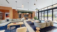 This thoughtfully designed communal space combines a grand lobby with a series of interconnected amenity areas, all unified by a cohesive, contemporary aesthetic. Polished terrazzo flooring extends throughout, reflecting natural light and lending an open, airy quality to every zone.

The entry lobby commands immediate attention with its soaring double-height ceiling, floor-to-ceiling glazing, and a monumental staircase framed by warm wood paneling. Curated artwork and lush potted plants punctuate the space, adding warmth and visual interest alongside a reception desk finished in natural wood tones.

Moving deeper into the space, a generously scaled lounge area features an expansive built-in shelving wall lined with books, décor, and trailing greenery set against a deep teal-painted ceiling. Varied seating arrangements—including plush sofas, sculptural lounge chairs, and a tufted daybed—invite both social gatherings and quiet relaxation. Pendant lighting in mixed styles adds character overhead, while large glazed doors offer views of an outdoor pool area.

A dedicated kitchen and gathering zone includes warm wood cabinetry, black countertops, a decorative patterned tile backsplash, open shelving adorned with plants, and a central bar-height communal table—ideal for casual meetings or social events.