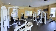 Located at 2100 Farm Springs Rd in Summerville, South Carolina, this single-floor fitness center offers a well-appointed workout space designed to support a comprehensive exercise routine. The gym is outfitted with a variety of strength-training machines, including multi-functional cable and weight systems by Hoist, as well as cardio equipment such as an elliptical and rowing machine, providing users with a full range of training options. The space features a dark rubber flooring throughout, ideal for absorbing impact and providing stability during workouts. Warm yellow walls paired with white crown molding give the room a clean, inviting aesthetic. Recessed lighting in the ceiling, complemented by a centrally mounted ceiling fan, ensures proper illumination and air circulation throughout the space. Multiple windows along the walls allow generous amounts of natural light to filter in, offering views of the surrounding greenery and creating an open, energizing atmosphere. The thoughtful layout allows ample room to move between equipment comfortably, making this fitness center a practical and welcoming environment for residents or members looking to maintain an active lifestyle.
