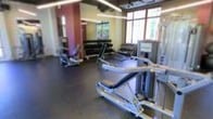 This fitness center is a fully equipped, single-floor facility designed to support a comprehensive workout experience. The space is outfitted with a diverse selection of strength training machines, including cable and resistance equipment, positioned thoughtfully throughout the floor to allow for comfortable movement between stations. A dedicated free weights area features a full rack of dumbbells alongside adjustable benches, catering to users of all fitness levels. The flooring is finished with durable, dark rubber matting, a practical and professional choice well-suited to high-traffic athletic use. Large windows along one wall allow an abundance of natural light to fill the space, creating a bright and energizing atmosphere that enhances the overall workout environment. Recessed linear lighting along the ceiling provides additional illumination, ensuring the space is well-lit at all times. Mirrors are strategically placed to give users clear sightlines for form correction and to visually expand the space. The layout is open and functional, maximizing usable floor space while maintaining clear pathways between equipment. The overall ambiance is clean, modern, and purpose-driven, making this fitness center a well-rounded facility for strength training and general fitness.