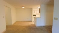 This single-floor residential unit offers a straightforward and functional layout designed for comfortable everyday living. The open-concept living and dining area is finished with neutral beige carpeting that creates a warm and inviting atmosphere, while the adjoining kitchen and entryway transition to durable tile flooring. The space is bathed in soft, even lighting from flush-mount ceiling fixtures, contributing to a clean and well-lit environment throughout.

The kitchen is partially open to the main living area, separated by a peninsula-style breakfast bar that serves as both a practical workspace and a casual dining surface. White cabinetry lines the kitchen walls, offering ample storage, while the countertops and appliances maintain the space's consistent neutral palette. An overhead range hood and stovetop are visible, rounding out the kitchen's functional amenities.

The living area is generously proportioned, featuring smooth white walls and standard-height ceilings that keep the space feeling open and airy. The overall design is simple and understated, making it highly versatile and easy to personalize. The clean lines, neutral finishes, and practical flow between spaces make this unit a functional and move-in-ready residence.