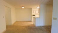 This single-floor residential unit offers a straightforward and functional layout designed for comfortable everyday living. The open-concept living and dining area is finished with neutral beige carpeting that creates a warm and inviting atmosphere, while the adjoining kitchen and entryway transition to durable tile flooring. The space is bathed in soft, even lighting from flush-mount ceiling fixtures, contributing to a clean and well-lit environment throughout.

The kitchen is partially open to the main living area, separated by a peninsula-style breakfast bar that serves as both a practical workspace and a casual dining surface. White cabinetry lines the kitchen walls, offering ample storage, while the countertops and appliances maintain the space's consistent neutral palette. An overhead range hood and stovetop are visible, rounding out the kitchen's functional amenities.

The living area is generously proportioned, featuring smooth white walls and standard-height ceilings that keep the space feeling open and airy. The overall design is simple and understated, making it highly versatile and easy to personalize. The clean lines, neutral finishes, and practical flow between spaces make this unit a functional and move-in-ready residence.