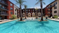 This outdoor pool area is designed to deliver a resort-like experience within a residential setting. The centerpiece is a generously sized swimming pool with vivid, crystal-clear blue water that reflects the open sky above. Tall palm trees are strategically positioned throughout the pool deck, providing a tropical aesthetic and natural shade during sunny days.

A raised, tiled platform at the pool's center serves as a distinctive architectural feature, flanked by swing-style loungers that add a playful and relaxed element to the space. Additional chaise lounge chairs are arranged along the pool's perimeter, offering ample spots for sunbathing and relaxation.

A covered pergola structure anchors one side of the pool area, outfitted with plush, white outdoor sectional seating and a coffee table, creating an ideal social gathering space. A mounted outdoor television and decorative lattice screen panel add functional and aesthetic value to this shaded retreat.

Adjacent to the main pool, a separate spa provides an additional relaxation option. The pool area is accessed through a dedicated entrance with rustic wood-framed doors set within a stucco exterior. The surrounding multi-story building architecture, featuring brick, stone, and contemporary design elements, frames the entire space, creating an enclosed and private outdoor oasis.