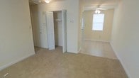 This single-floor residential space presents a straightforward and functional layout finished in a soft, neutral color palette throughout. The interior is characterized by wall-to-wall carpeting that runs continuously across the rooms, lending a cohesive and comfortable feel underfoot. A prominent feature of the space is the generously sized closet, fitted with bifold louvered doors and an interior rod and shelf system, providing practical and organized storage. The ceiling fan with a built-in light fixture is a notable detail, ensuring both air circulation and overhead illumination. Adjacent rooms connect openly, allowing natural light from the windows to filter through and brighten the interior. The walls are finished in a clean, light cream tone, complementing the neutral carpeting and white trim work, including visible baseboards that frame each room neatly. Additional doors suggest further rooms or utility access, indicating a multi-room floor plan. Overall, the space conveys a clean, move-in-ready character with simple, classic finishes suited to a variety of furnishing styles. The layout is practical, with attention given to storage and comfortable living areas on a single, accessible level.
