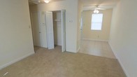 This single-floor residential space presents a straightforward and functional layout finished in a soft, neutral color palette throughout. The interior is characterized by wall-to-wall carpeting that runs continuously across the rooms, lending a cohesive and comfortable feel underfoot. A prominent feature of the space is the generously sized closet, fitted with bifold louvered doors and an interior rod and shelf system, providing practical and organized storage. The ceiling fan with a built-in light fixture is a notable detail, ensuring both air circulation and overhead illumination. Adjacent rooms connect openly, allowing natural light from the windows to filter through and brighten the interior. The walls are finished in a clean, light cream tone, complementing the neutral carpeting and white trim work, including visible baseboards that frame each room neatly. Additional doors suggest further rooms or utility access, indicating a multi-room floor plan. Overall, the space conveys a clean, move-in-ready character with simple, classic finishes suited to a variety of furnishing styles. The layout is practical, with attention given to storage and comfortable living areas on a single, accessible level.