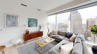 This high-rise residence is defined by its contemporary design and exceptional city views, visible through expansive floor-to-ceiling windows that span multiple rooms. The open-concept living area serves as the centerpiece of the space, featuring light hardwood flooring, crisp white walls, and generous natural light. A large sectional sofa anchors the living room, which flows seamlessly toward a corner window arrangement offering panoramic urban vistas.

The kitchen is sleek and well-appointed, combining white gloss upper cabinetry with light wood lower cabinets and a chevron-patterned tile backsplash. Stainless steel appliances, a linear pendant light, and a freestanding island with bar seating complete the space.

The residence includes multiple bedrooms, each thoughtfully designed with light flooring and neutral tones. The primary bedroom benefits from large windows with striking skyline views, while a secondary bedroom offers a quieter, more intimate setting. Both rooms feature clean lines and ample light.

Two bathrooms serve the residence: one with a glass-enclosed walk-in shower and wood-toned vanity cabinetry, and a second featuring a full soaking tub with shower combination and large-format tile surround. Together, the spaces offer a cohesive, modern aesthetic throughout.