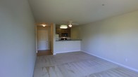 This single-floor residential unit offers a well-organized, open-concept layout designed for comfortable everyday living. The main living area is generously proportioned, featuring light-colored wall-to-wall carpeting that creates a warm, inviting atmosphere. A ceiling fan with an integrated light fixture is centrally positioned overhead, providing both airflow and ambient illumination throughout the space.

The kitchen is seamlessly integrated into the living area via a raised peninsula counter, which serves as a natural divider while maintaining the open feel of the floor plan. Dark wood cabinetry lines the kitchen walls, complemented by a built-in microwave and refrigerator, offering practical storage and appliance options. The contrasting dark countertops add a grounded aesthetic touch to the space.

A hallway visible from the main living area leads to the entry door, which features a classic paneled design. The transition from carpeted living space to hard flooring in the entryway corridor is clearly defined, contributing to a functional flow through the unit. Neutral-toned walls throughout the space provide a versatile backdrop, making the interior easy to personalize. Overall, the unit presents a clean, practical, and cohesive living environment suited to a range of lifestyles.