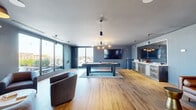 This contemporary amenity lounge is a thoughtfully designed social hub spread across a single level, offering a variety of distinct zones for entertainment and leisure. The main game room is anchored by a full-sized pool table set atop a teal area rug, with warm hardwood flooring throughout and slate-blue accent walls that lend a modern, cohesive aesthetic. A large wall-mounted flat-screen TV and a well-appointed wet bar with stainless steel appliances complete this space, while floor-to-ceiling windows flood the room with natural light and frame open city views.

Adjacent to the game room, a comfortable lounge area features plush barrel chairs and accent tables, with large sliding glass doors leading directly to an outdoor terrace. The terrace is outfitted with string lights, planters, pergola structures, and communal seating, creating an inviting open-air extension of the indoor space.

A dedicated dining area offers a mix of high-top tables with upholstered chairs alongside an elegant tufted banquette with marble-top tables beneath warm pendant lighting and a decorative wall mural. The overall ambiance throughout is refined yet relaxed, combining contemporary furnishings, curated artwork, and abundant natural light to create an exceptional shared environment suited for both casual gatherings and more intimate social occasions.