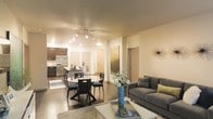 This single-floor space showcases an open-concept layout that fluidly connects the kitchen, dining, and living areas into one cohesive environment. The kitchen is equipped with dark wood cabinetry, stainless steel appliances including a full-size refrigerator, and a central island that serves as both a workspace and a casual gathering point. Track lighting runs along the ceiling, casting warm, directed light across the space and complementing the neutral-toned walls.

The dining area is positioned centrally, anchored by a round table with sleek modern chairs, creating an inviting setting for meals and conversation. Polished concrete floors extend throughout, lending an industrial yet refined character to the interior. The living area features a contemporary grey sofa accented with decorative pillows in muted tones, paired with a glass coffee table and a textured area rug that defines the seating zone. Decorative touches, including a tall blue vase with floral stems and wall art, add personality to the space. The overall ambiance is modern and well-lit, with a cohesive design aesthetic maintained throughout.