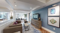 This thoughtfully designed single-floor space showcases a seamless open-plan layout that connects multiple functional zones through consistent architectural detailing. Throughout the space, warm-toned herringbone hardwood flooring creates a striking visual foundation, complemented by white coffered ceilings with crown molding and recessed lighting that add a refined, classical character.

The living area features a bold slate-blue accent wall housing a wall-mounted television above a sculptural geometric media console. Comfortable neutral seating is arranged around a layered blue area rug, with the space opening fluidly toward a dining area. Structural white columns with wainscoting panels elegantly define zones without interrupting sightlines.

The dining area is bathed in natural light through large windows dressed with striped drapery, centered beneath a modern circular pendant light. A generously sized walnut dining table with contemporary upholstered chairs anchors the room, accompanied by a statement indoor plant.

A well-appointed wet bar includes dark cabinetry, a mosaic tile backsplash, open floating shelves, a built-in stainless microwave, sink, and a wine refrigerator. A separate lounge nook with patterned wallpaper and a glass-paneled partition adds a private retreat within the open floor plan.
