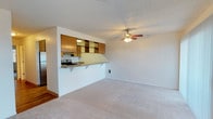 This single-floor space offers a practical and comfortable layout designed to maximize everyday functionality. The open-concept living area is finished with plush wall-to-wall carpeting and bright white walls, creating a clean and inviting atmosphere. A textured ceiling adds a classic touch throughout, while a ceiling fan with a built-in light fixture provides both comfort and ambient lighting to the main living space.

The kitchen is positioned adjacent to the living area and is separated by a raised breakfast bar that doubles as a casual dining counter, encouraging an easy flow between spaces. Warm wood-toned upper cabinetry with mirrored cabinet faces lines the kitchen wall, offering generous storage while adding a reflective quality that brightens the area. The kitchen is equipped with an electric range and a sink, providing all the essential amenities for meal preparation. An under-cabinet range hood is installed above the stovetop for ventilation.

A wall-mounted air vent is visible in the living area, ensuring climate control throughout the space. The transition from carpet to hardwood-style flooring near the kitchen entry adds visual variety and a sense of defined zones within the open layout. Overall, the space presents a straightforward, well-maintained interior with functional design elements suited for comfortable single-floor living.