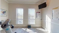 This fitness center is designed to provide a comprehensive workout environment within a single, well-organized space. The room features light wood-look flooring throughout, crisp white wainscoting along the lower walls, and neutral warm-toned upper walls, creating a clean and welcoming aesthetic. Crown molding adds a refined architectural detail, lending the space a more polished feel than a typical gym setting.

Natural light fills the room through multiple tall windows fitted with white horizontal blinds, allowing adjustable light control throughout the day. A wall-mounted flat-screen television is positioned for visibility across the workout area, providing entertainment during exercise sessions.

The fitness center is equipped with a treadmill, recumbent stationary bike, and an elliptical-style machine, offering a solid range of cardiovascular options. Kettlebells and a stability ball are also available for functional and strength training. Exercise mats provide dedicated space for floor-based workouts. Multiple workout reference posters — covering kettlebell routines, bodyweight exercises, suspension training, and muscle group guides — are displayed on the walls, serving as useful visual aids.

A glass-panel door connects the fitness center to an adjacent hallway, contributing to an open, accessible feel. Overall, the space is bright, functional, and thoughtfully arranged for a variety of fitness activities.