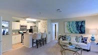 This single-floor residence offers an open-concept layout that connects the kitchen and living area in a seamless, functional design. The kitchen is outfitted with stainless steel appliances, white cabinetry, and a granite-topped peninsula with stylish upholstered bar stools — ideal for casual dining or entertaining. Adjacent to the kitchen, the living area is finished with plush carpeting and soft, neutral wall tones that create a calm, inviting atmosphere. Coastal-inspired décor, including a striking blue abstract artwork, reinforces the light and airy feel throughout the space. A doorway off the main living area leads to additional rooms, suggesting a practical and well-organized floor plan. The textured ceiling adds subtle architectural character, while centrally placed lighting and HVAC vents contribute to a comfortable interior environment. Natural light enhances the open feel of the space, highlighting the cohesive color palette of whites, soft greens, and blues. Overall, this residence combines modern conveniences with a relaxed, welcoming ambiance, making it a comfortable and visually appealing living space.