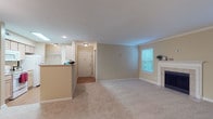 This single-floor residence is thoughtfully laid out to maximize functionality across its living spaces. The living room serves as the central gathering area, finished with plush wall-to-wall carpeting, crown molding, and a classic white-mantled fireplace with tile surround. Two windows fitted with blinds allow natural light to brighten the room while maintaining privacy.

The kitchen is designed in a galley configuration, lined on both sides with light wood cabinetry and ample counter space. White appliances, including a range, microwave, and refrigerator, are neatly integrated. A peninsula counter separates the kitchen from the main living area, creating a natural transition between the two spaces. Recessed lighting and hardwood-style flooring complete the kitchen's practical aesthetic. Adjacent to the kitchen, a dedicated laundry area provides added convenience.

The bedroom is a well-proportioned, carpeted space featuring a ceiling fan with integrated lighting and a door that opens directly to the exterior. The full bathroom includes hardwood-style flooring, a tub and shower combination with a curtain rod, a vanity with under-cabinet storage, a large mirror, and pendant lighting overhead. Warm, neutral tones are consistent throughout the entire unit, creating a cohesive and understated atmosphere.