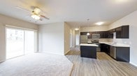 This single-floor residence offers a seamlessly connected living and kitchen layout, ideal for both everyday living and entertaining. The living area is finished with plush carpet and features a ceiling fan, creating a comfortable and relaxed atmosphere. Natural light filters in through a sliding glass door, brightening the space and adding an airy feel. The transition from carpet to wood-look flooring marks the entry into the kitchen and dining area, providing a clean visual distinction between spaces. The kitchen is well-appointed with dark espresso cabinetry that contrasts elegantly against light countertops, offering generous storage and workspace. A central island serves as a functional hub for meal preparation and casual dining. Stainless steel appliances, including a refrigerator, microwave, and range, add a modern, polished touch to the culinary space. Pendant lighting above the island provides focused illumination while enhancing the kitchen's contemporary aesthetic. A hallway visible from the main living area leads to additional rooms, suggesting a thoughtfully designed floor plan. The neutral color palette throughout — soft beige walls and light flooring — creates a cohesive, welcoming environment adaptable to a variety of interior styles.