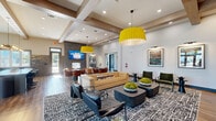 Located at 3145 Prado Ln in Charlotte, NC, this expansive single-floor amenity facility offers a thoughtfully designed collection of spaces built for comfort, recreation, and community. The welcoming lounge area makes an immediate impression with its soaring ceilings adorned with exposed wood beam detailing, bold yellow pendant lighting, rich hardwood floors, and a full built-in shelving wall with integrated media display. Curated furnishings, patterned area rugs, and framed artwork contribute to a polished yet inviting atmosphere. A communal kitchen nearby features sage green cabinetry with brass hardware, a dramatic textured brick backsplash, a large center island, stainless steel appliances, and an artisan globe pendant light cluster — ideal for gatherings and events. The fitness center is generously sized, flooded with natural light through large windows, and outfitted with a comprehensive range of cardio and strength equipment, ceiling fans, and wall-mounted displays. An adjoining functional training room provides space for dedicated workout routines with outdoor views. The golf simulator room, finished in dark tones with turf flooring and a ceiling-mounted projector, offers an immersive experience. The game room rounds out the amenities with a billiards table, lounge seating, wainscoting detail, and a wall-mounted TV — perfect for relaxed socializing.