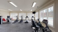 Located on Ligon Mill Road in Wake Forest, NC, this single-story fitness center is designed to support a comprehensive workout experience across multiple interconnected zones. The facility is thoughtfully organized to separate cardio and strength training areas, creating a functional and efficient layout.

The cardio area is bright and airy, lined with large windows fitted with white blinds that invite generous natural light and views of the surrounding greenery. This space houses a selection of treadmills, elliptical trainers, and recumbent bikes, all arranged along the windowed walls. A wall-mounted television adds convenience for users during their sessions. Overhead, flush-mounted fluorescent lighting ensures consistent illumination throughout.

A dedicated strength training zone features a robust collection of equipment, including cable machines, weight benches, a dumbbell rack, and selectorized machines with red upholstered seating. Crown molding and neutral warm-toned walls lend a polished, well-maintained appearance to this area. French-style glass panel doors connect the zones, enhancing the sense of openness.

An additional room accommodates functional fitness tools such as stability balls, an air resistance bike, and a weight bench, offering space for cross-training and flexibility work. Dark rubber flooring throughout provides durability and traction across all areas.