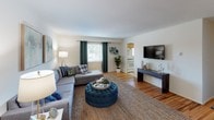 This single-floor residence is thoughtfully arranged to maximize functionality across its well-defined living spaces. The living room serves as the central gathering area, featuring warm hardwood flooring, large windows dressed with teal curtains that invite natural light, and ample room for comfortable seating arrangements. A wall-mounted television and open sightlines into the kitchen make this space ideal for everyday living.

The adjacent kitchen and dining area are practical and well-equipped, featuring warm wood cabinetry, a gas range, dishwasher, and a window above the sink that provides natural light and outdoor views. A dedicated dining space within the kitchen accommodates a full dining table, making it suitable for both casual meals and entertaining.

The unit includes multiple bedrooms, each finished with plush carpeting and neutral walls. The primary bedroom is notably spacious, featuring multiple windows, generous closet space, and room for a sitting area. Additional bedrooms feature hardwood flooring, individual closets, and windows for natural light.

The bathroom is finished with classic tile wainscoting, a single-sink vanity with wood cabinetry, a well-lit mirror, and patterned tile flooring, contributing a clean and timeless aesthetic to the space overall.