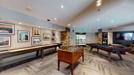 Located at 3202 W Colfax Ave in Denver, Colorado, this single-floor amenity lounge is a thoughtfully designed social and recreational space that blends modern sophistication with warm, rustic-industrial character. Reclaimed wood accent walls and stone-clad columns serve as defining architectural elements throughout, creating a cohesive and inviting atmosphere.

The game room area is well-appointed with a regulation billiards table, a full-length shuffleboard table, and a foosball table, all set atop herringbone-patterned carpet tile flooring. Framed urban photography prints of Denver cityscapes line the walls, adding a locally inspired aesthetic touch. Recessed lighting illuminates the space effectively throughout.

Adjacent to the game area, a stylish wet bar features a dramatic black granite waterfall-edge countertop, complemented by sleek bar stools with brass-toned legs. Teardrop pendant lights hang above the bar, while rustic wood planking frames a built-in display with dual wall-mounted screens and a subway tile backsplash, creating a refined yet casual atmosphere.

The lounge seating area offers tufted leather sectional sofas arranged around cylindrical gold accent tables, all positioned in front of a large wall-mounted screen set against a deep teal accent wall. Flanking built-in bookshelves with curated décor complete the space, making it ideal for relaxed gatherings or media viewing.