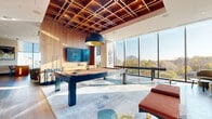 This upscale amenity floor is thoughtfully designed to deliver a premium social and recreational experience. The centerpiece game room impresses with a striking coffered wood ceiling, warm walnut wall paneling, and a large-format mounted display. A full-size billiards table anchors the space atop an artful area rug, complemented by a shuffleboard table and plush leather seating. Floor-to-ceiling windows flood the room with natural light and frame lush treetop views. A dedicated golf simulator room offers a high-tech Full Swing projection system, acoustic wall panels, and a turf hitting mat for an immersive experience. The main lounge area features expansive glazing with panoramic city views, wide-plank hardwood flooring, and an eclectic mix of modern seating — including rounded armchairs in warm gold and neutral tones — creating an inviting, light-filled gathering space. A built-in bar with open shelving and a wine refrigerator adds convenience for entertaining. A separate lounge nook provides a more intimate retreat, furnished with a deep-seated modular velvet sofa, curated abstract wall art, and soft, layered lighting. Together, these spaces combine refined materials, contemporary design, and versatile programming to create an exceptional amenity environment.