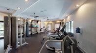 This fitness center is a thoroughly outfitted exercise facility designed to support a wide variety of workout routines. The space features durable rubber flooring throughout, tray ceilings with recessed lighting, and ceiling fans that maintain air circulation across the workout floor. Large windows and glass doors along one wall provide views of an outdoor pool area while allowing natural light to fill the interior.

The cardio zone includes multiple treadmills, a spin bike, and an elliptical machine, all positioned to take advantage of the outdoor views. A wall-mounted flat-screen television adds an entertainment option for cardio sessions. On the opposite end of the facility, a dedicated strength and functional training area houses a multi-station cable machine, a heavy punching bag, resistance bands, and a wall-mounted storage rack stocked with medicine balls and additional accessories. A full dumbbell rack with a broad range of free weights anchors the center of the space, complemented by a weight bench for targeted exercises. Separate restroom facilities are accessible directly from the gym floor, and a secondary entrance connects to the outdoors. The overall layout is open and well-organized, providing clear zones for cardio, strength training, and functional fitness without congestion.